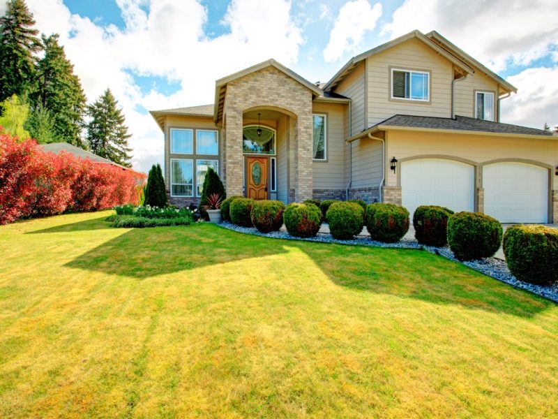 Big siding house with garage and high column porch. Green lawn with trimmed hedges and red bushes make the curb appeal stand out